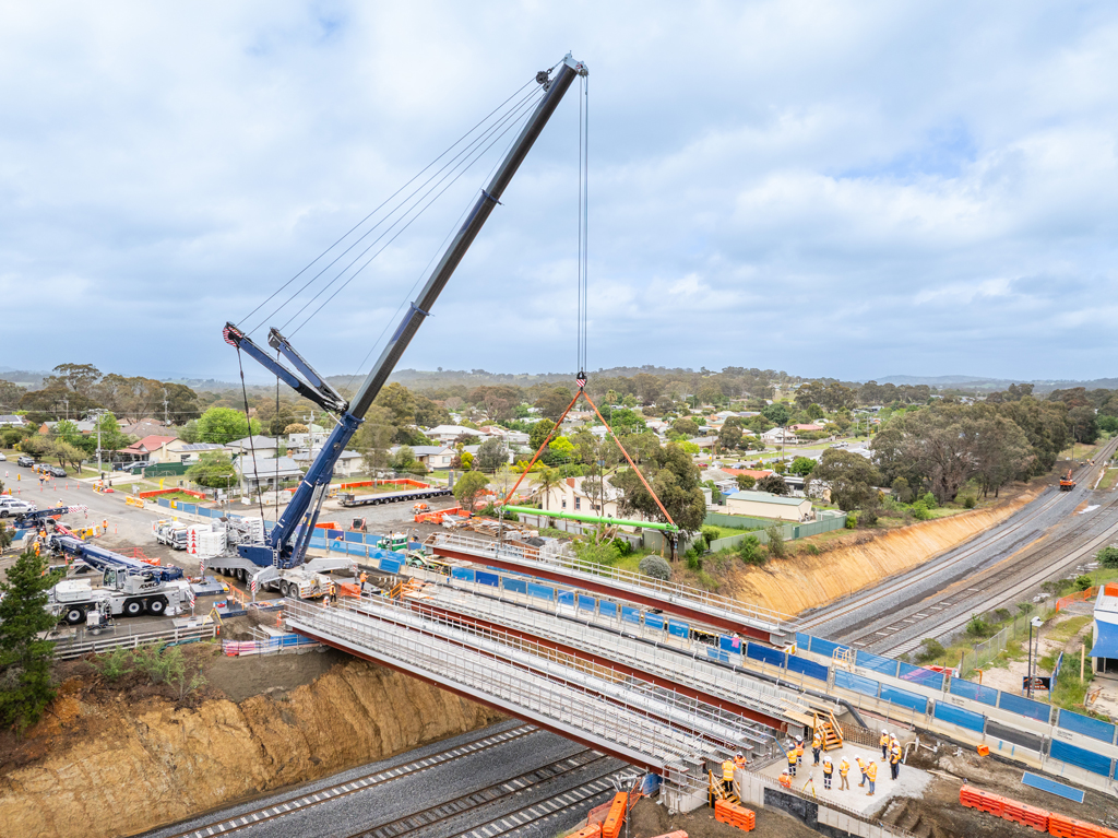 A crane lifts a bridge structure into place over a road and rail bridge, in an urban setting.