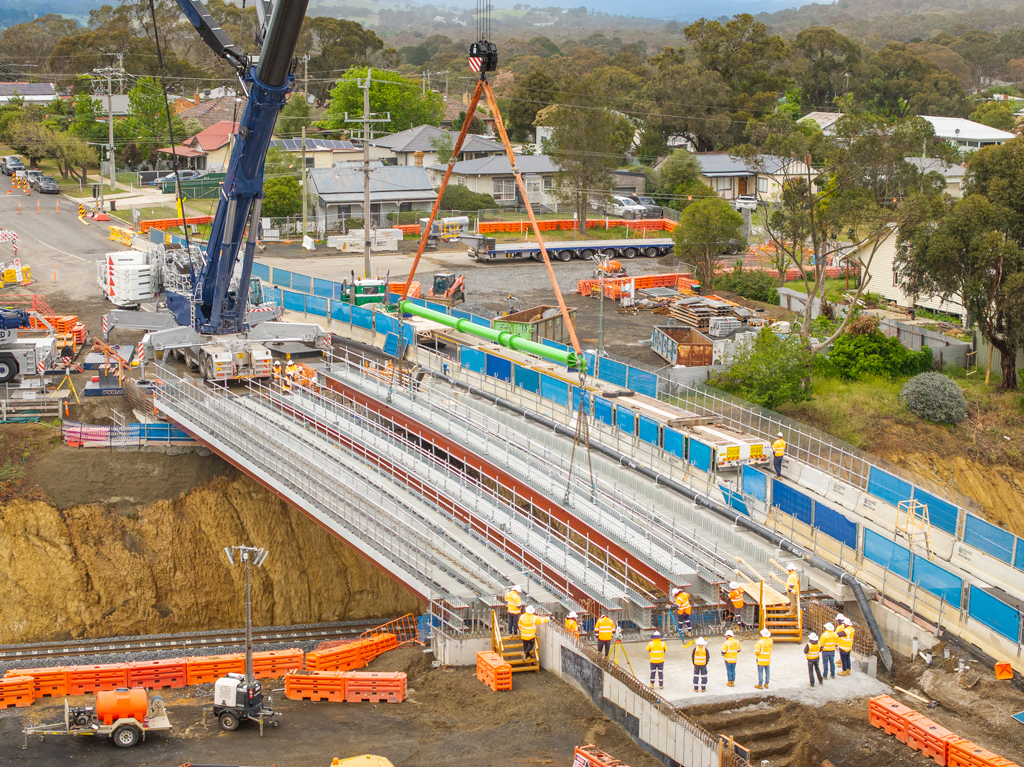 A crane lifts a bridge structure into place over a road and rail bridge, in an urban setting.