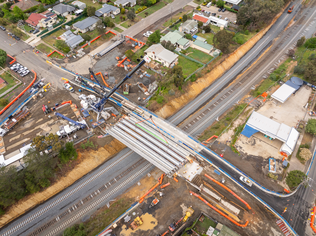 A crane lifts a bridge structure into place over a road and rail bridge, in an urban setting.