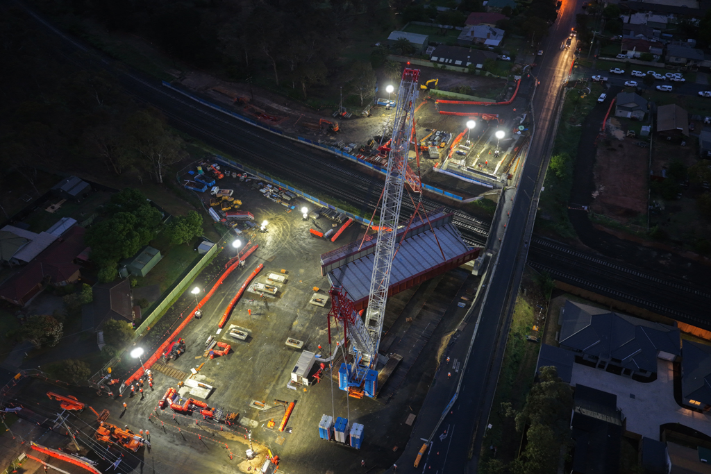 A crane lifts a bridge structure into place over a road and rail bridge, in an urban setting.