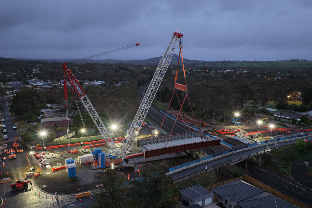 A crane lifts a bridge structure into place over a road and rail bridge, in an urban setting.