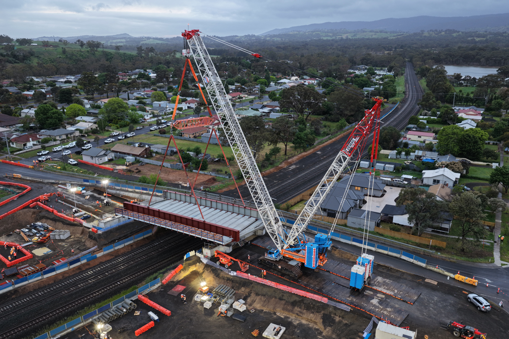 A crane lifts a bridge structure into place over a road and rail bridge, in an urban setting.