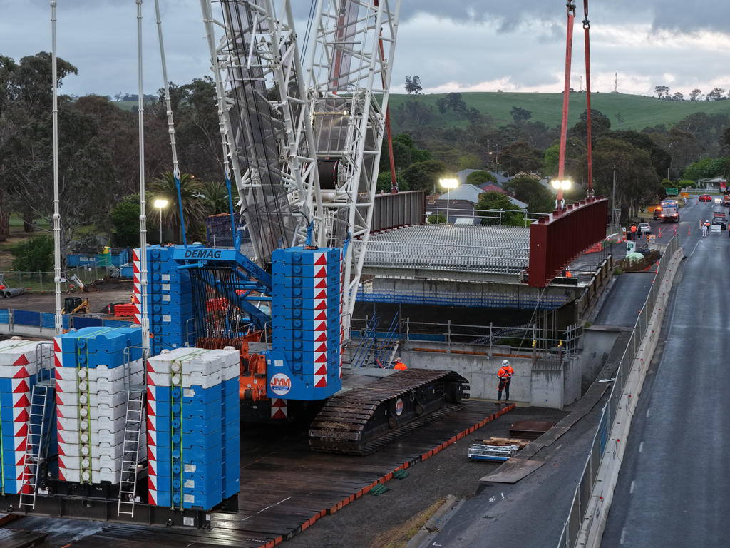 A crane lifts a bridge structure into place over a road and rail bridge, in an urban setting.