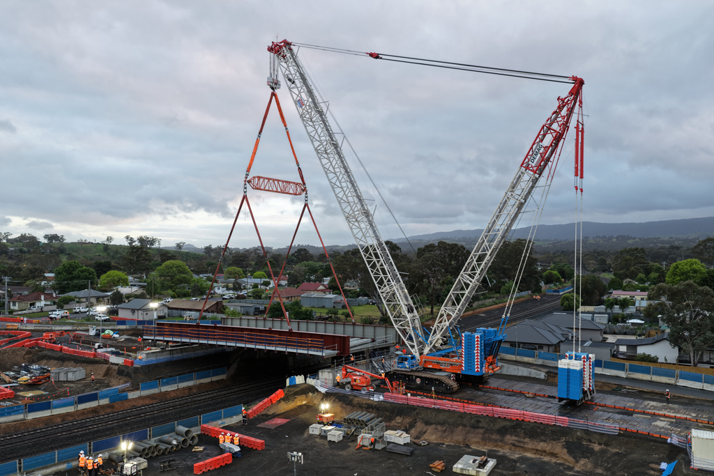 A crane lifts a bridge structure into place over a road and rail bridge, in an urban setting.