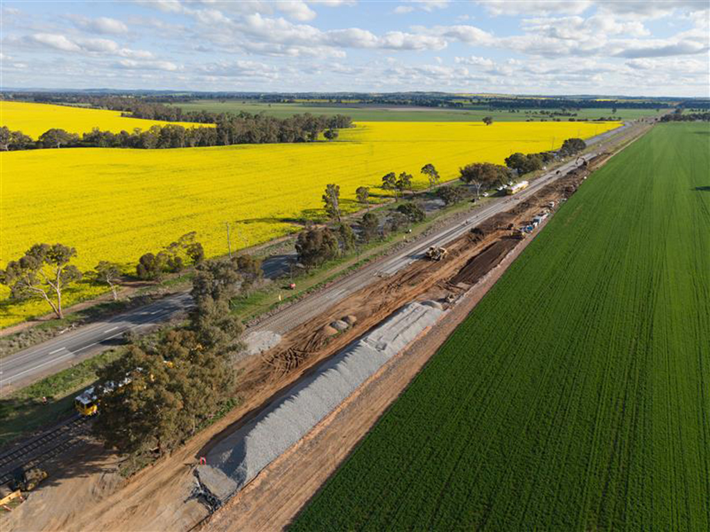 Aerial view of a railway construction site and adjacent road cutting through farmland with green and yllow crops.