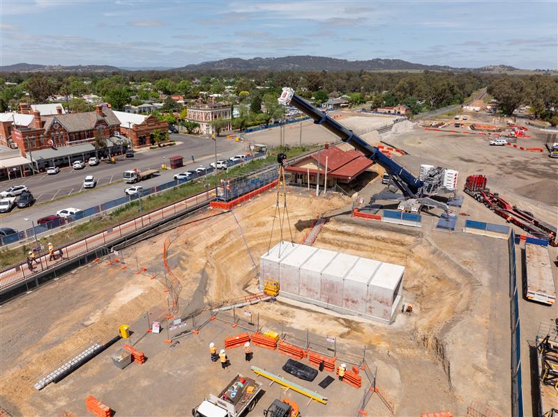 Construction site with a large concrete structure being lowered into an excavated area by a crane near a town street.