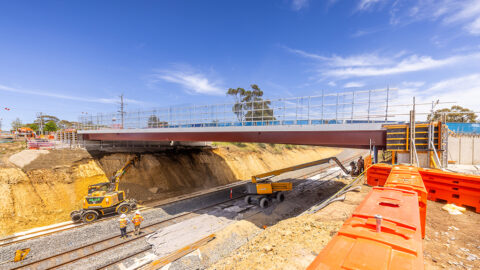 Construction site showing a newly built road bridge over train tracks, with excavator and dump truck working below and orange safety barriers in the foreground.