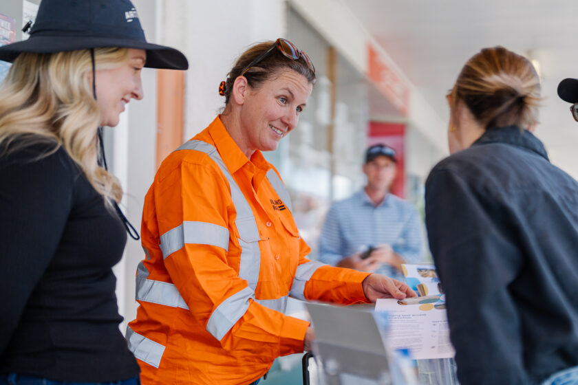 Two people in uniforms for Inland Rail talk to another person outside a row of shops.