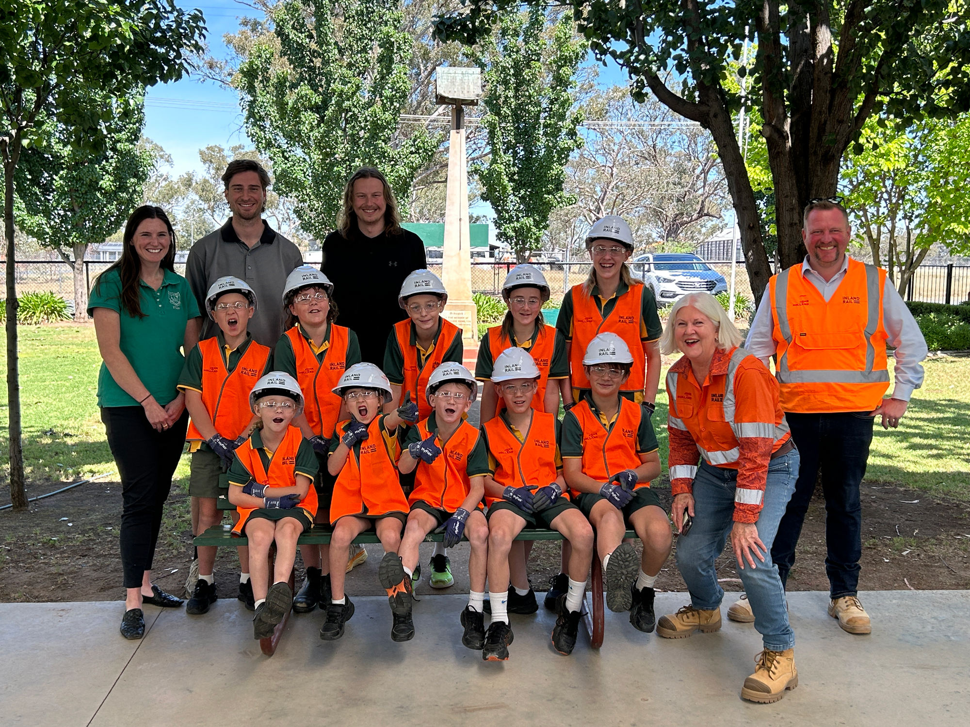 A number of students lined up in two rows wear high visibility safety vests and hard hats, with adults standing nearby, in a classroom-style photo outside.