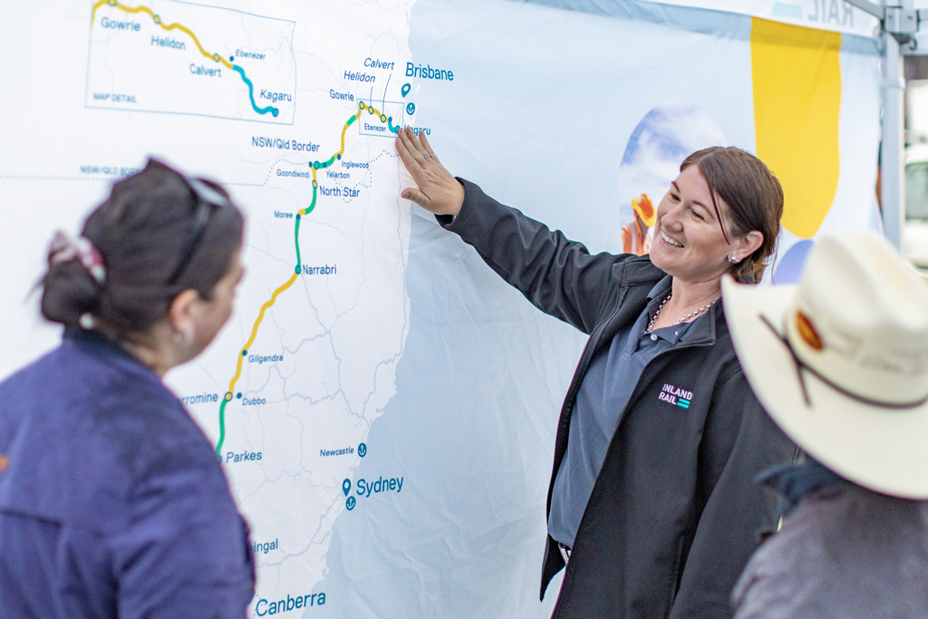 A person stands in front of a large map, pointing to a position on it, while talking to two other people.