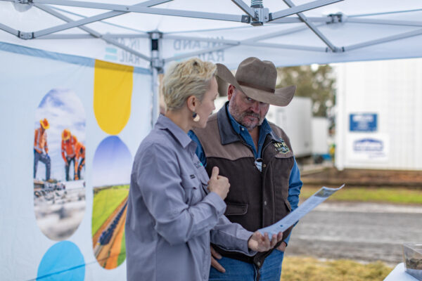 Two people stand at an information booth at a show. One person wears and Inland Rail uniform, and holds a piece of paper, explaining information to the other person, who is wearing a wide-brimmed hat.