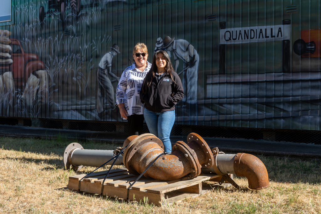Two people stand behind a pile of metal pipes, in front of a heritage mural with the name Quandialla pictured in the mural.