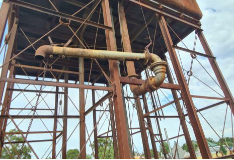 A metal structure on stilts, with a water pipe attached to the side of it. All the metal is covered in rust. In a rural setting.
