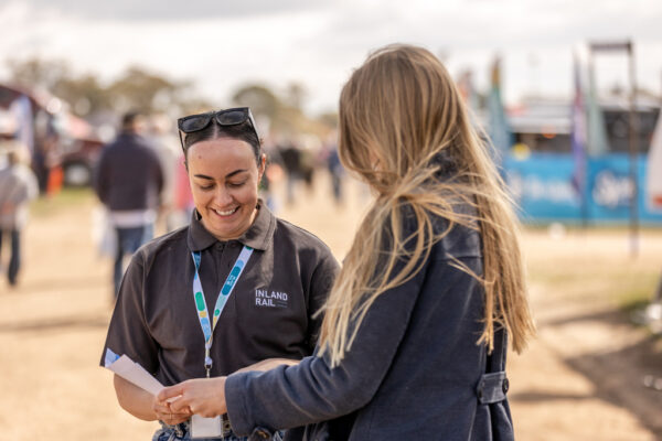 Two people stand in the outdoors. One wears and Inland Rail uniform and lanyard, and is explaining information on a sheet to another person who is also holding the sheet of paper.