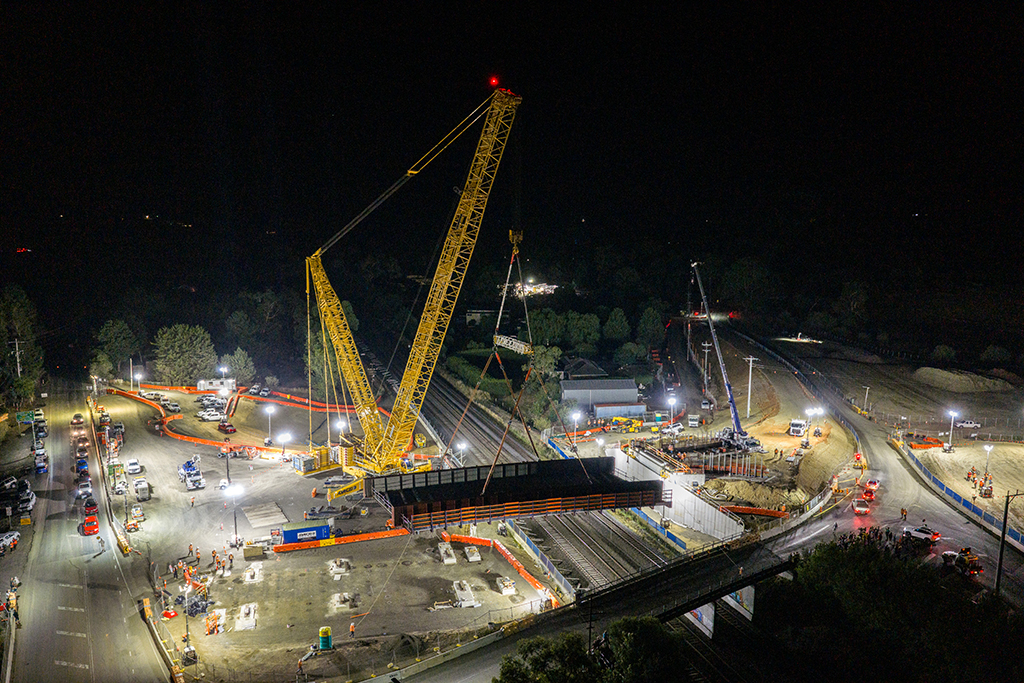 Aerial night view of cranes installing a bridge section over railway tracks, surrounded by construction activity.