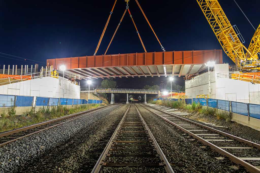 A large steel bridge section being lifted into place over railway tracks at night using cranes.