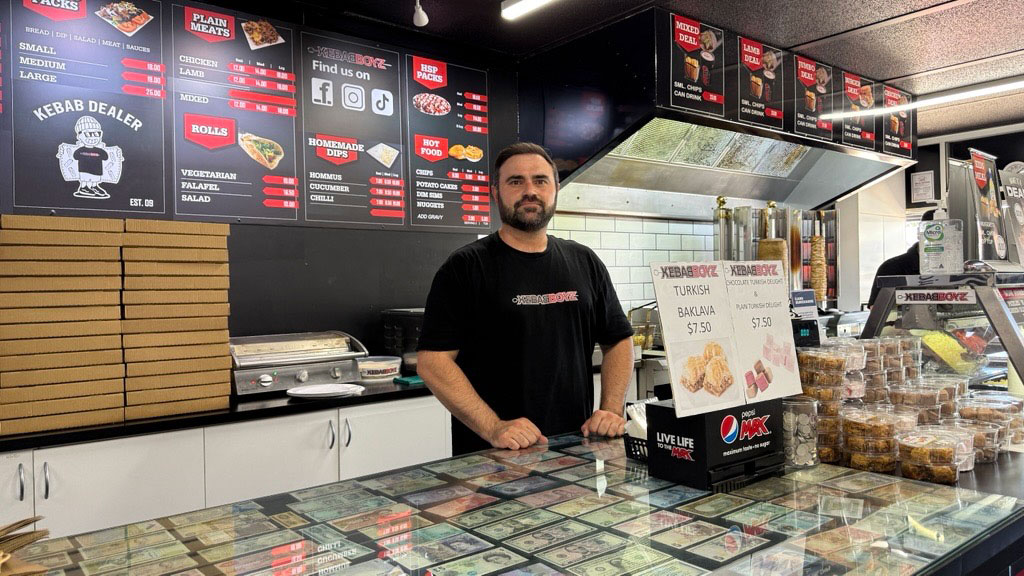 Owner standing behind the counter inside a kebab shop with menu boards and food displays visible.