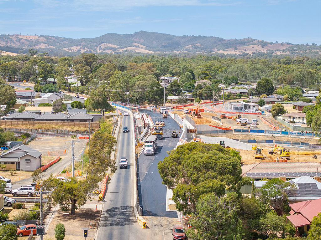 Aerial view of a road bridge construction site through a residential area, with traffic moving past earthworks and machinery.
