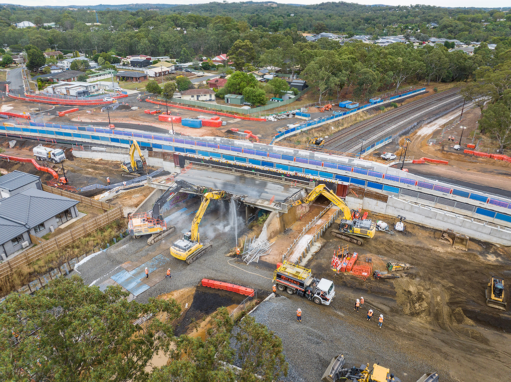 Aerial view of bridge construction over rail tracks with excavators and workers below.