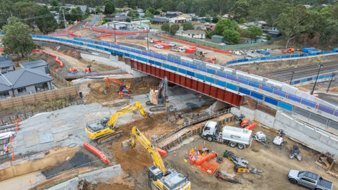 Aerial view of bridge construction over rail tracks with excavators and workers below.
