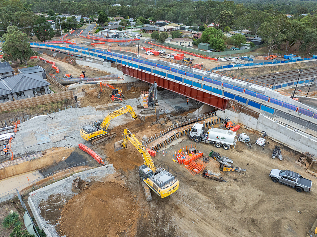 Aerial view of bridge construction over rail tracks with excavators and workers below.
