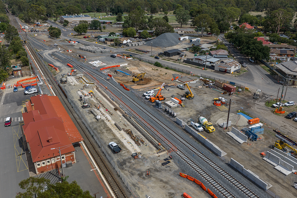 Aerial view of rail tracks and station area with construction works, machinery and safety barriers along the corridor.