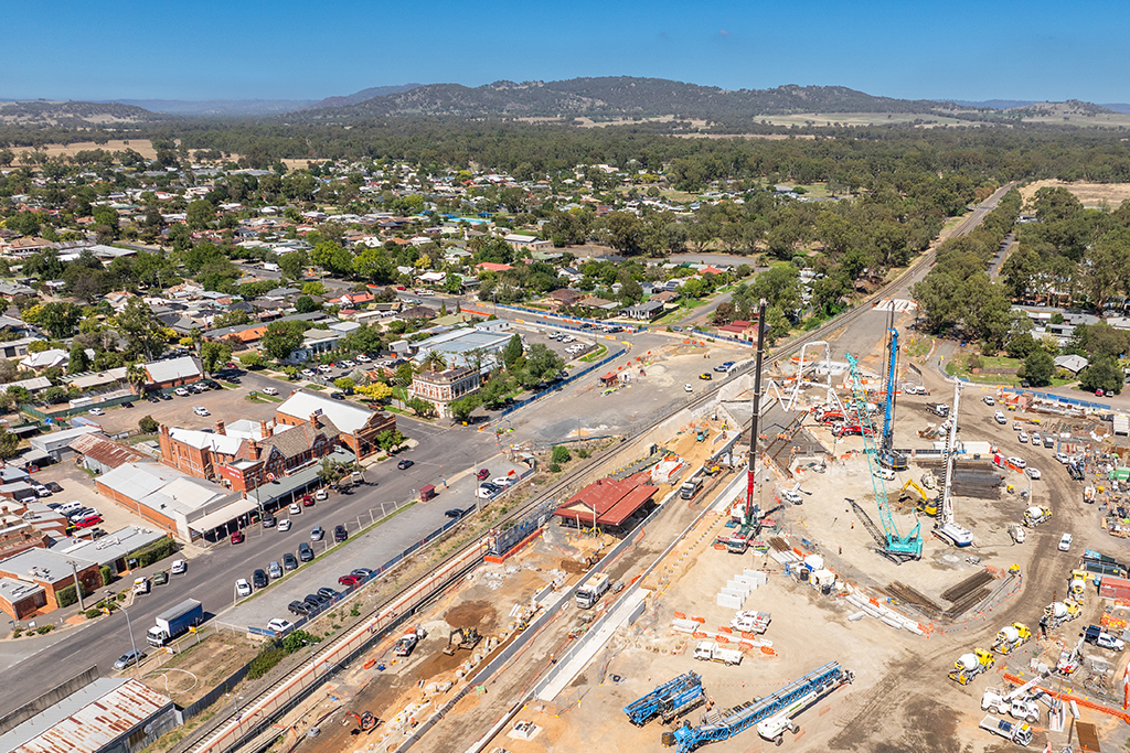 Aerial view of a town rail corridor with station works underway, including cranes, machinery and concrete construction.