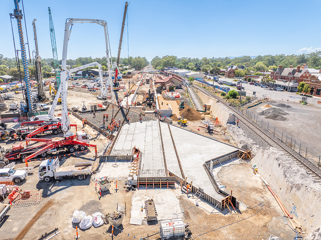 Aerial view of concrete works underway at a rail station, with cranes, trucks, workers and rail tracks visible.