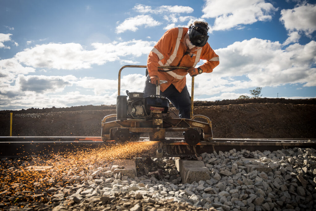 A worker dressed in safety gear uses a machine to cut track next to a railway line. Sparks fly from the bottom of the machine.