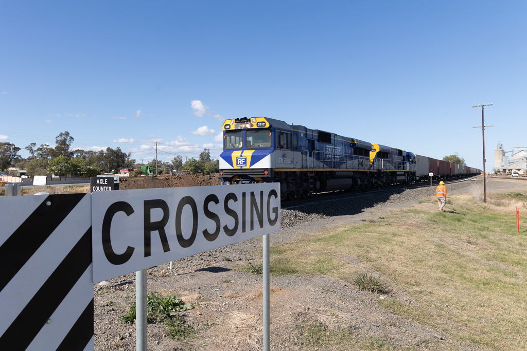 A freight train approaches a crossing with a silo and industrial site in the distance, in a rural area. A worker walks down the embankment.