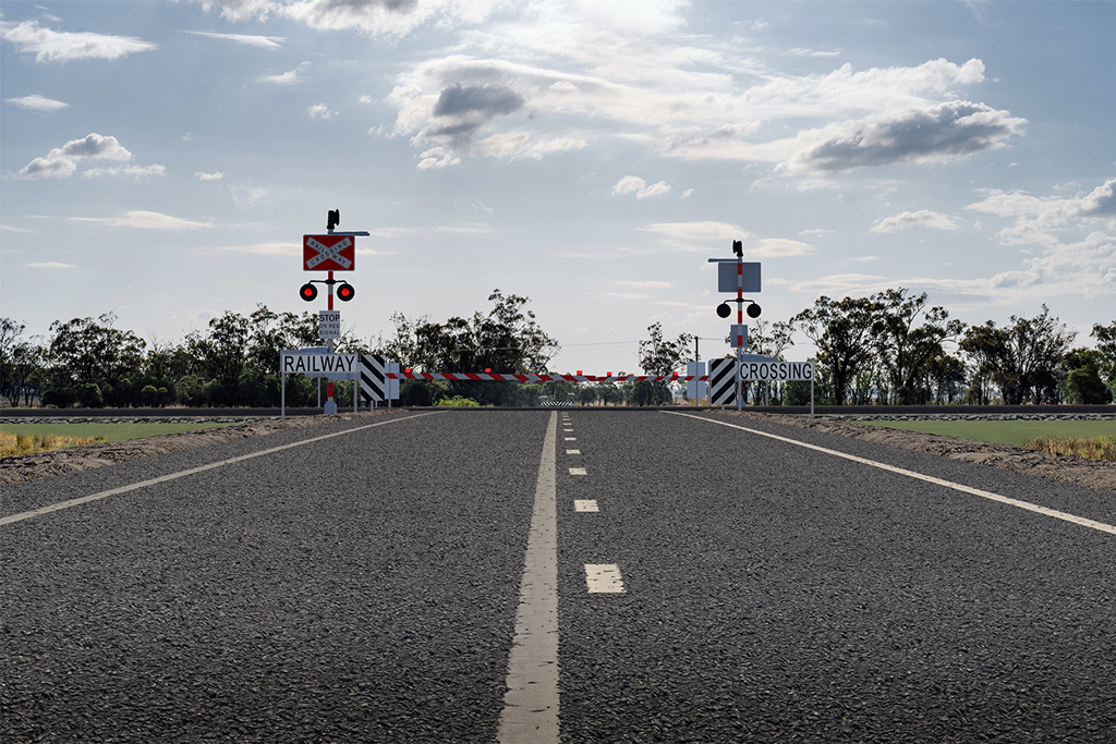 A rural road leads to an intersection in a rural area. A railway crosses the road, and a level crossing signage and slights can be seen before each side of the railway facing oncoming traffic.