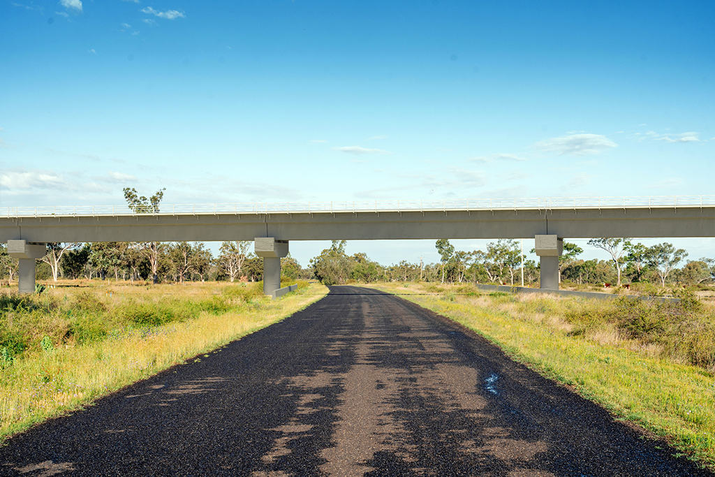 A rural scene showing a bitumen road, flanked by grass and trees, curving off into the distance. A rail bridge extends over the road from one side of the frame to the other.