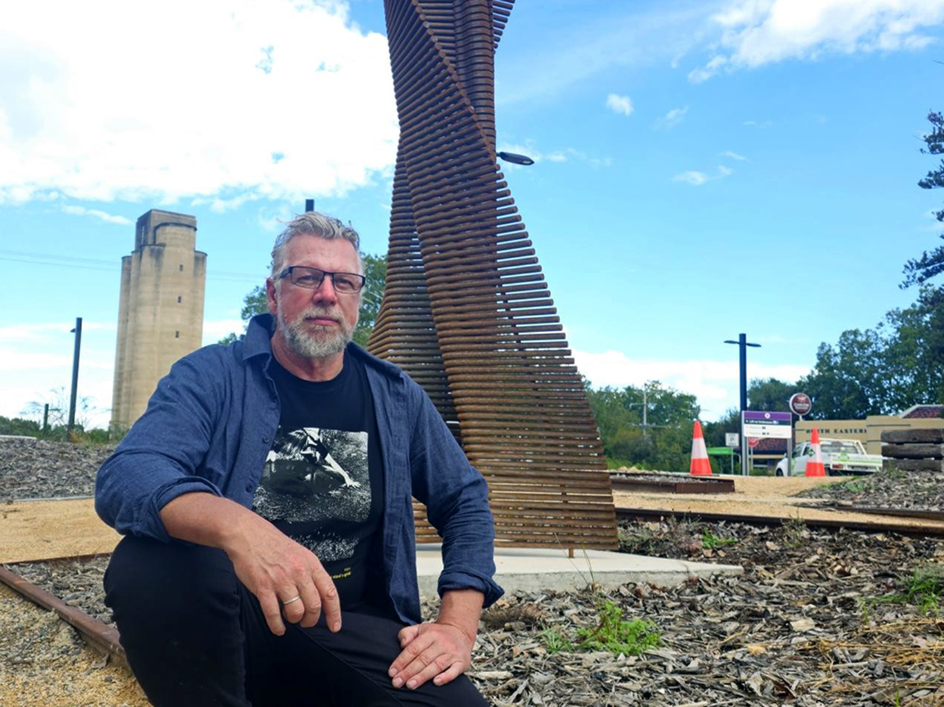 Artist seated beside a timber sculpture near a rail line, with station buildings and tracks in the background.