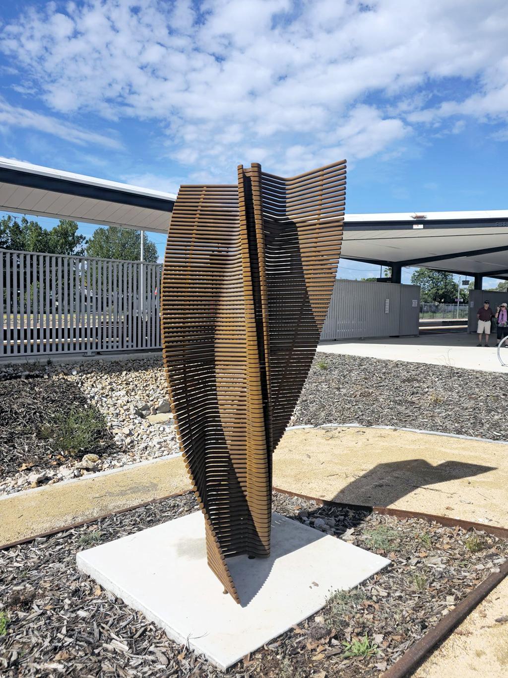 Timber sculpture installed on a concrete base near a station platform and pedestrian area.