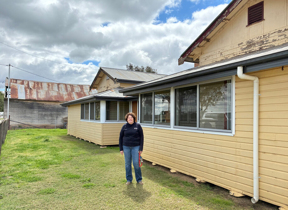 A woman stands in front of a house in a rural landscape.