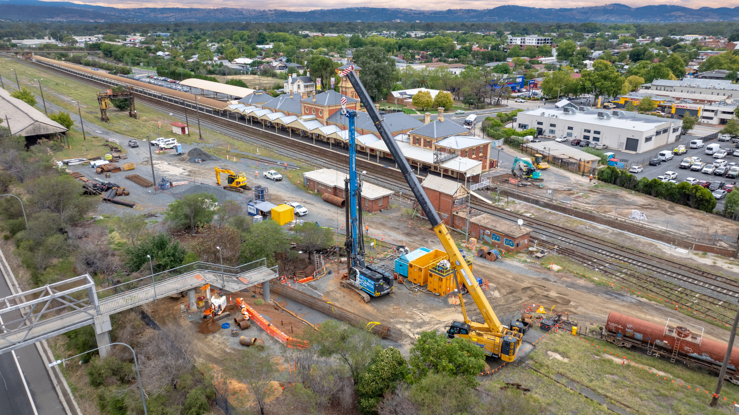 A crane and a pile driving machine at a worksite beside a railway track and railway station in a regional town.