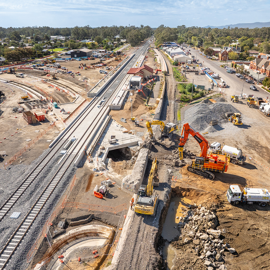 A busy worksite with three heavy vehicles digging next to a railway track. A train station is in the centre of the work, in a regional town.