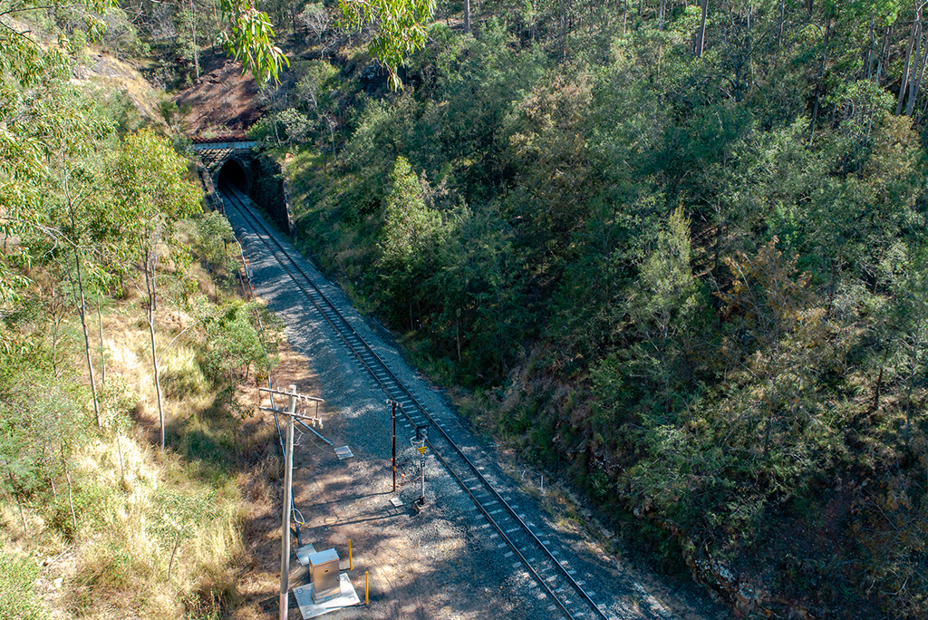 In a valley between two treed bills, a railway line stretches into a tunnel under a mountain.