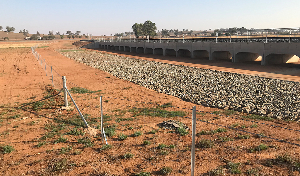 A wore fence extends alongside a culvert with a railway track running over it. It is in a rural field, with red dirt and a farm complex in the background.