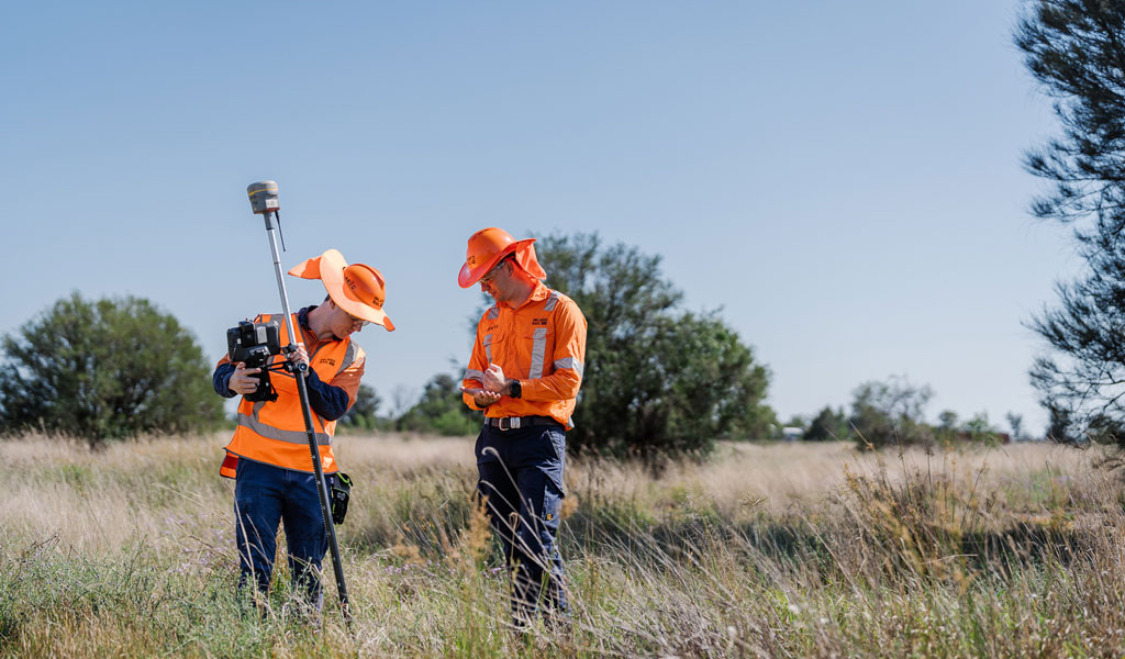 Two people in high visibility workwear stand in a field. One is holding a pile with scientific equipment. The other is jotting something down on an electronic tablet.