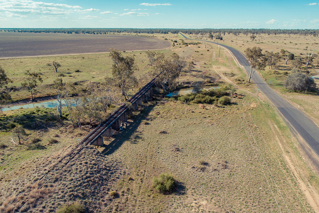 An old railway line runs parallel to a road in a rural area, with farmland surrounding them.