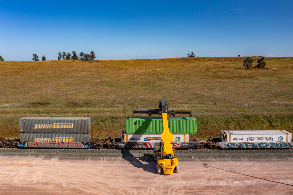 A shipping container is stacked on top of a train that already has one container on it, in a rural field. 