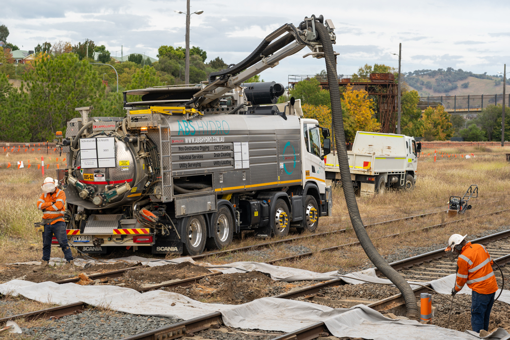 A truck with a tube attached to it pumps something into the ground next to a railway line that is under construction. 