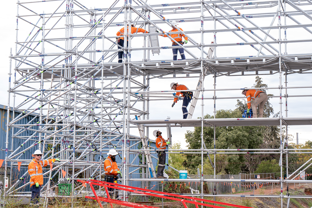 A scaffolding bridge stretches over a railway line. Workers are at various heights on the scaffold, constructing it.