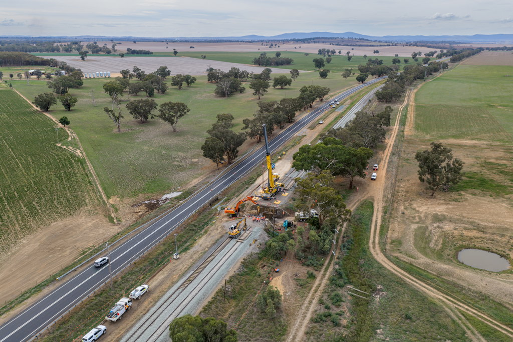 A railway line stretches across a rural landscape. A work crew uses heavy equipment to adjust the track on a bridge.