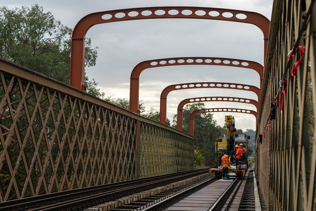 Workers stand on a railway bridge. The bridge has railings in a criss-cross pattern. A series of steel arches connect the two sides of the railings.