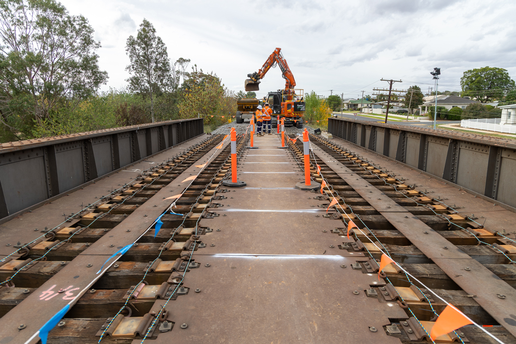 Work crews watch as heavy machinery moves soil just past a steel railway bridge in a urban area.
