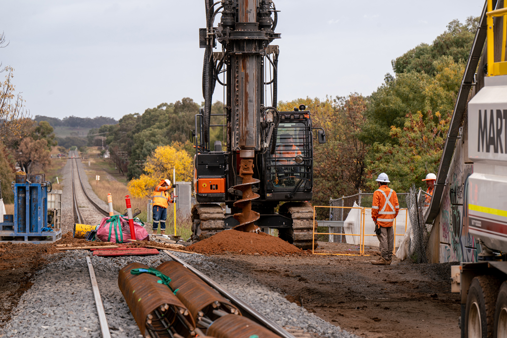 A large drill machine digs a hole next to a railway track in a worksite under a bridge.