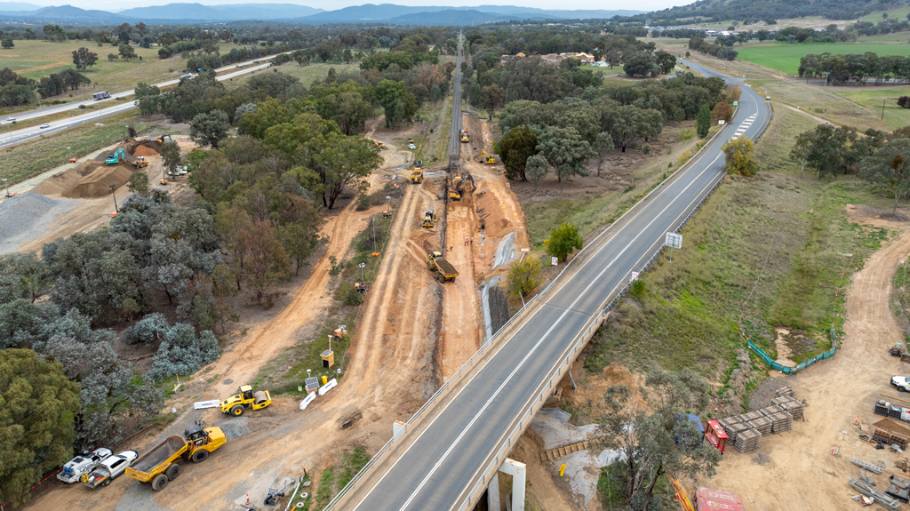 A road bridge continues over a railway worksite. The railway line stretches to the horizon in this rural landscape.