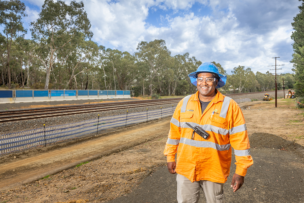 Worker in high‑visibility clothing and a hard hat standing beside railway tracks at a construction site.